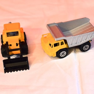 May include: Two toy construction vehicles: a yellow front-end loader with a black bucket and a yellow dump truck with a silver bed. Both have black wheels and are on a white surface.