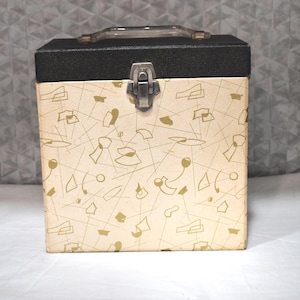 May include: A square storage case with a black textured lid and a cream-colored body featuring a gold-toned abstract geometric pattern. The case includes a clear plastic top handle and a central silver-toned metal latch closure on the front.