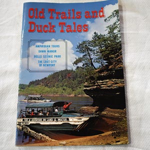 Op de afbeelding: Een vintage reisbrochure getiteld Old Trails and Duck Tales met een foto van een amfibievoertuig op een rivier bij rotsachtige kliffen. De tekst vermeldt Amphibian Tours, Dawn Manor, Dells Scenic Park en The Lost City of Newport.