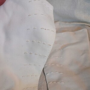 May include: Two white fabric pieces with rows of stitching. The fabric is folded and the stitching is visible on the edges.