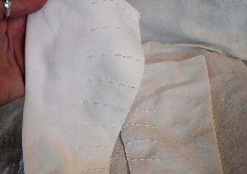 May include: Two white fabric pieces with rows of stitching. The fabric is folded and the stitching is visible on the edges.