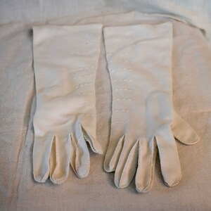May include: A pair of white long gloves with stitching detail. The gloves are made of a soft, smooth fabric.