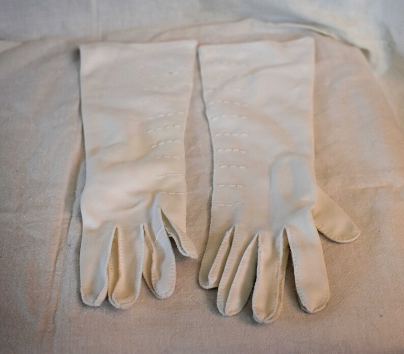 May include: A pair of white long gloves with stitching detail. The gloves are made of a soft, smooth fabric.