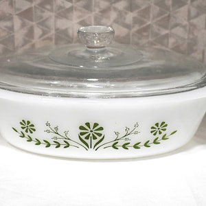 Peut inclure: Un plat à four ovale blanc en verre opalin avec un couvercle en verre transparent. L'extérieur présente un motif floral vert répétitif composé de marguerites stylisées et de vignes feuillues centrées autour d'une fleur plus grande.