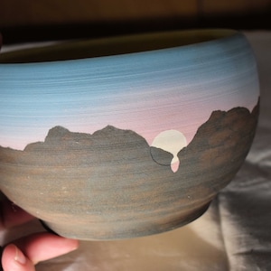 Peut inclure: Un bol en céramique avec une scène de coucher de soleil peinte. Le bol est brun avec un ciel bleu et rose et un soleil blanc. Les montagnes sont en silhouette contre le ciel.