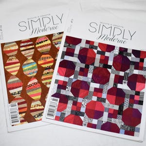 Puede incluir: Dos números de la revista Simply Moderne Quilts & Crafts, el número 10 y el número 7. La portada izquierda presenta un fondo marrón con bloques de edredón a rayas en forma ovalada. La derecha muestra un patrón geométrico en rojo, rosa y negro.