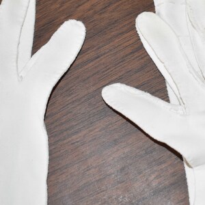 May include: A pair of white cotton gloves with a seam down the middle of each finger. The gloves are laid flat on a brown wooden surface.
