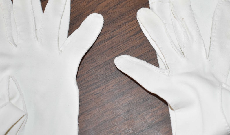 May include: A pair of white cotton gloves with a seam down the middle of each finger. The gloves are laid flat on a brown wooden surface.