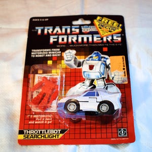 May include: Vintage Transformers Throttlebot Searchlight toy, still in its original packaging. The packaging features a red and black design with the Transformers logo and the text "Transforms from Motorized Minicar to Robot and Back!"