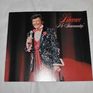 May include: A black and white photo of Liberace, a famous entertainer, wearing a sequined jacket and a red vest. The text "Liberace" and "Mr. Showmanship" is printed on the image.