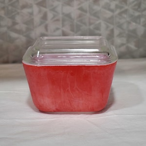 Puede incluir: Un recipiente cuadrado de vidrio rojo con una tapa de vidrio transparente. El recipiente tiene un aspecto ligeramente texturizado y se encuentra sobre una superficie blanca. La tapa es rectangular y encaja perfectamente.