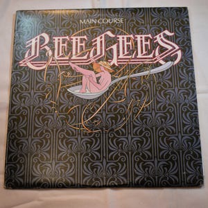 May include: Album cover for the Bee Gees' "Main Course." The cover features the band's name in a stylized font with a nude figure in a spoon. The background is a dark blue pattern with floral designs.