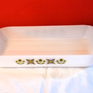 May include: White rectangular baking dish with green and gold floral pattern. The dish has handles on each end and is sitting on a white surface. The background is red.