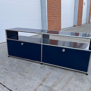 May include: A navy blue and silver modular storage unit with two sections. Each section has a dark blue door with a silver knob and a silver frame. The top shelf is silver and the unit is on a concrete surface.
