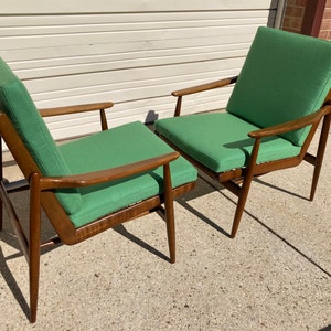 May include: Two mid-century modern chairs with wooden frames and green fabric upholstery. The chairs are positioned side-by-side, with the back of one chair facing the front of the other.