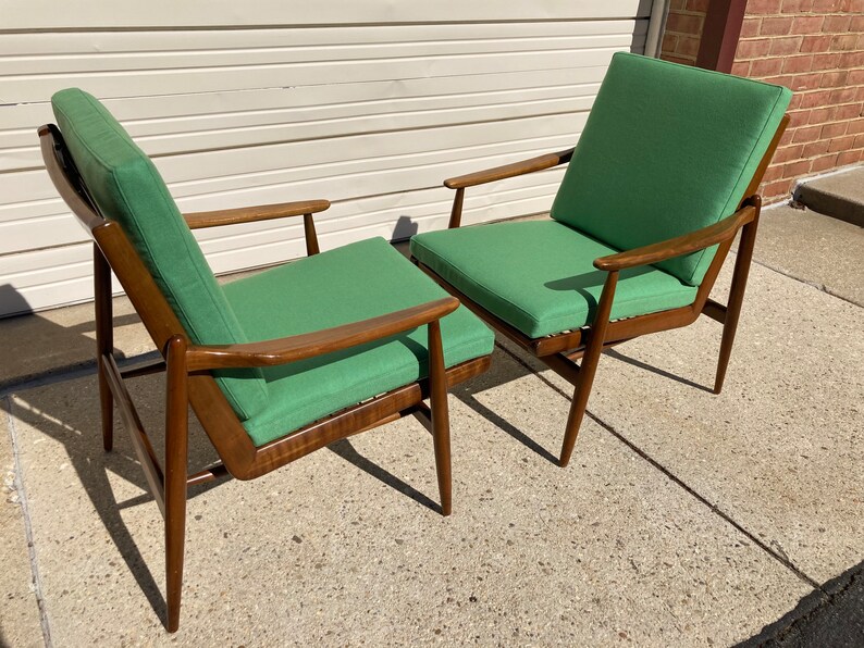 May include: Two mid-century modern chairs with wooden frames and green fabric upholstery. The chairs are positioned side-by-side, with the back of one chair facing the front of the other.