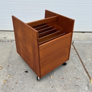 May include: A rectangular wooden storage unit with a warm brown finish, featuring multiple vertical interior dividers for organizing items. The unit is constructed with solid side panels and is supported by four small wheels attached to the base.