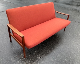 May include: A mid-century modern sofa with a red fabric upholstery and wooden frame. The sofa has a simple design with clean lines and a low profile.