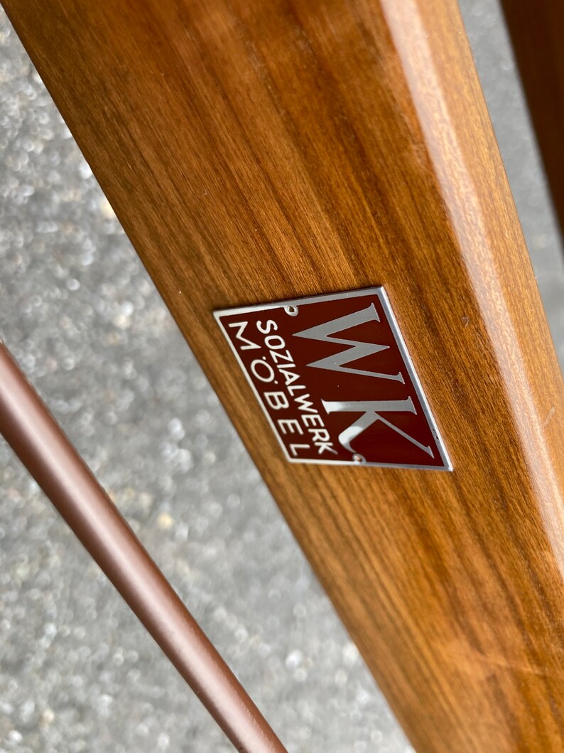 May include: A close-up of a wooden surface with a silver and red metal label that reads "WK Sozialwerk M&ouml;bel".