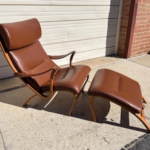 May include: A brown leather lounge chair with a matching ottoman. The chair has a high back and curved wooden arms. The ottoman is rectangular and has a curved wooden frame.