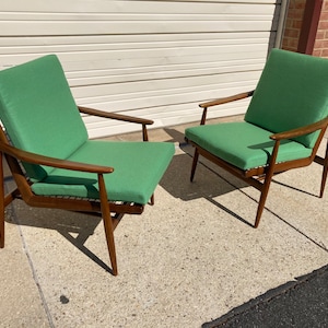 May include: Two mid-century modern chairs with a dark wood frame and green fabric upholstery. The chairs are sitting on a concrete floor.