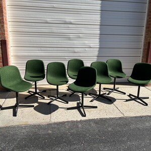May include: Eight green chairs with black metal legs and a swivel base. The chairs are arranged in a row on a concrete surface.