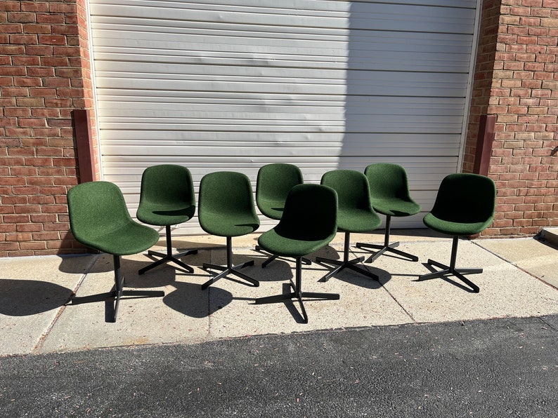 May include: Eight green chairs with black metal legs and a swivel base. The chairs are arranged in a row on a concrete surface.