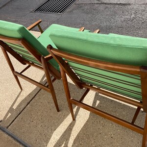 May include: Two mid-century modern chairs with a green fabric upholstery and a wooden frame. The chairs are angled and have a slatted back.