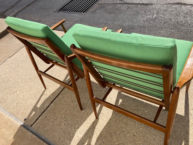May include: Two mid-century modern chairs with a green fabric upholstery and a wooden frame. The chairs are angled and have a slatted back.