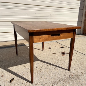 May include: A mid-century modern end table with a single drawer and tapered legs. The table is made of dark wood and has a simple, elegant design.