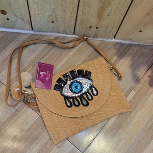 May include: A tan crossbody bag with a large blue eye with black eyelashes and white detailing. The bag has a woven texture and a small pom-pom charm.