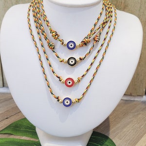 May include: Four layered necklaces with colorful woven cords and gold beads. Each necklace features a different colored evil eye charm with a white center and gold rim. The charms are blue, black, red, and blue.
