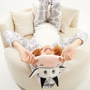 May include: A person wearing a gray and white giraffe-patterned onesie with a cow-themed hood and eye mask. The person is lying on a cream-colored chair with their legs in the air. The eye mask is pink and white.