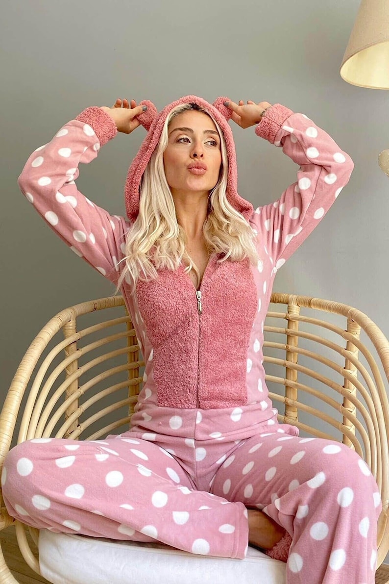 May include: A pink and white polka dot hooded onesie with bear ears. The onesie is made of a soft, plush fabric and has a zipper closure.