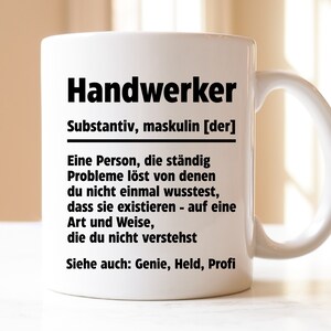 Könnte beinhalten: Weiße Keramiktasse mit schwarzem Design, das das Wort "Handwerker" als eine Person definiert, die Probleme auf einzigartige Weise löst. Der Text lautet: "Substantiv, maskulin [der] Eine Person, die ständig Probleme löst von denen du nicht einmal wusstest, dass sie existieren - auf eine Art und Weise, die du nicht verstehst. Siehe auch: Genie, Held, Profi"