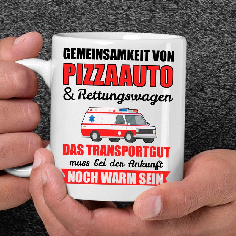 Puede incluir: Taza de cer&aacute;mica blanca con un dise&ntilde;o en negro y rojo. La taza presenta una ambulancia de dibujos animados con el texto "GEMEINSAMKEIT VON PIZZAAUTO & Rettungswagen" y "DAS TRANSPORTGUT muss bei der Ankunft NOCH WARM SEIN".