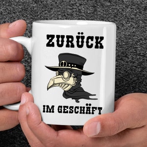 Könnte beinhalten: Weiße Keramiktasse mit einer schwarz-weißen Illustration eines Pestarztes mit Maske und Hut. Der Text "ZURÜCK IM GESCHÄFT" ist auf die Tasse gedruckt.