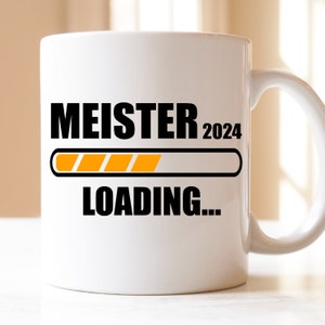 Könnte beinhalten: Weiße Keramik-Kaffeetasse mit schwarzem Text, der "MEISTER 2024" und "LOADING..." lautet, mit einer Fortschrittsanzeige darunter. Die Fortschrittsanzeige ist mit Orange gefüllt.