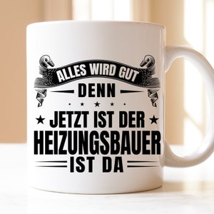 Könnte beinhalten: Weiße Keramiktasse mit schwarzem Text, der "Alles wird gut denn jetzt ist der Heizungsbauer ist da" lautet.