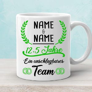 Könnte beinhalten: Weiße Keramiktasse mit grünen Lorbeerkränzen und schwarzem Text, der "NAME & NAME 12,5 Jahre Ein unschlagbares Team" lautet, mit zwei schwarzen und weißen ineinander verschlungenen Ringen.