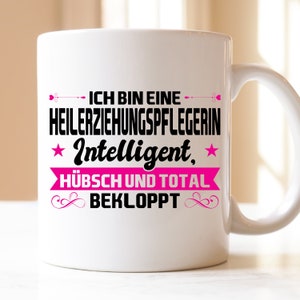 Könnte beinhalten: Weiße Keramiktasse mit einem schwarzen und pinkfarbenen Design. Der Text auf der Tasse lautet "Ich bin eine Heilerziehungspflegerin Intelligent, hübsch und total bekloppt".