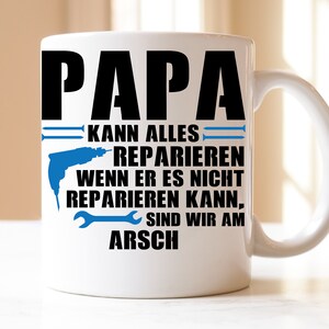 Könnte beinhalten: Weiße Keramiktasse mit einem schwarzen und blauen Design. Die Tasse zeigt den Text "PAPA KANN ALLES REPARIEREN WENN ER ES NICHT REPARIEREN KANN, SIND WIR AM ARSCH".