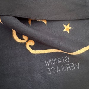 Op de afbeelding: Een zwarte zijden sjaal met een gouden geborduurd ontwerp met een ster en de tekst "VERSACE GIANNI".