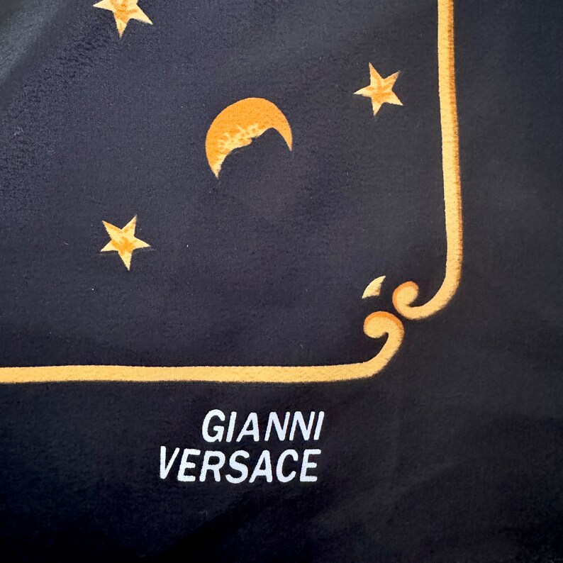 Op de afbeelding: Zwarte zijden sjaal met een gouden rand en een ontwerp met een halve maan en sterren. De sjaal heeft de tekst "GIANNI VERSACE" in wit gedrukt.