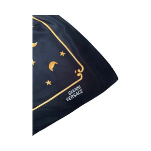 Op de afbeelding: Zwarte zijden sjaal met een gouden geborduurde rand en een maan- en sterrendesign. De sjaal heeft de tekst "GIANNI VERSACE" in wit geborduurd.
