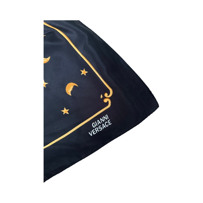 Op de afbeelding: Zwarte zijden sjaal met een gouden geborduurde rand en een maan- en sterrendesign. De sjaal heeft de tekst "GIANNI VERSACE" in wit geborduurd.