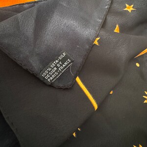 Op de afbeelding: Een zwarte zijden sjaal met een gouden sterrenpatroon en de tekst "100% seta-silk design by Paris-France" op een label.