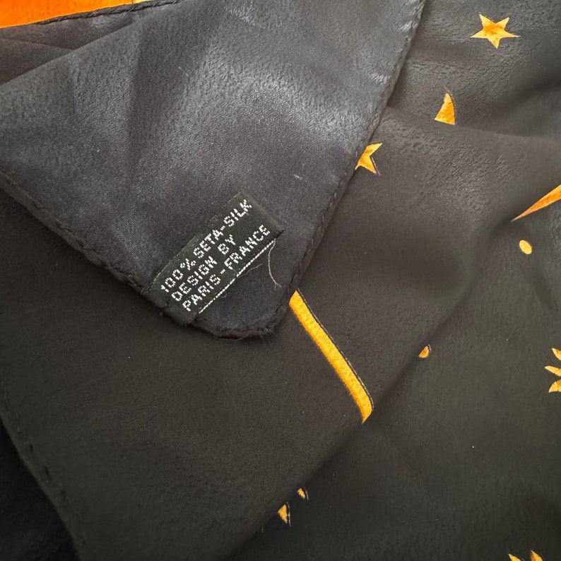 Op de afbeelding: Een zwarte zijden sjaal met een gouden sterrenpatroon en de tekst "100% seta-silk design by Paris-France" op een label.