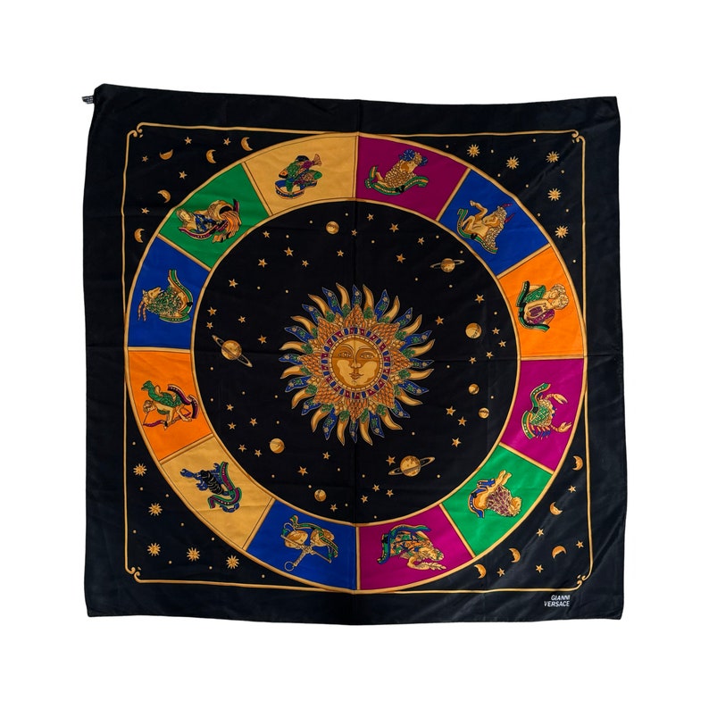 Op de afbeelding: Een zwarte vierkante sjaal met een kleurrijk ontwerp van een dierenriemwiel. Het wiel toont de 12 dierenriemtekens, elk in een andere kleur, en een zon in het midden. De sjaal is afgewerkt met een gouden rand en heeft de tekst "Gianni Versace" in de rechterbenedenhoek.