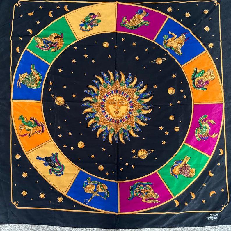 Op de afbeelding: Een zwarte vierkante zijden sjaal met een gouden zodiac-ontwerp. Het midden van de sjaal heeft een gouden zon met een gezicht. De dierenriemtekens zijn in een cirkel rond de zon afgebeeld. De sjaal is afgewerkt met een gouden rand en heeft de tekst "Gianni Versace" in de rechterbenedenhoek.